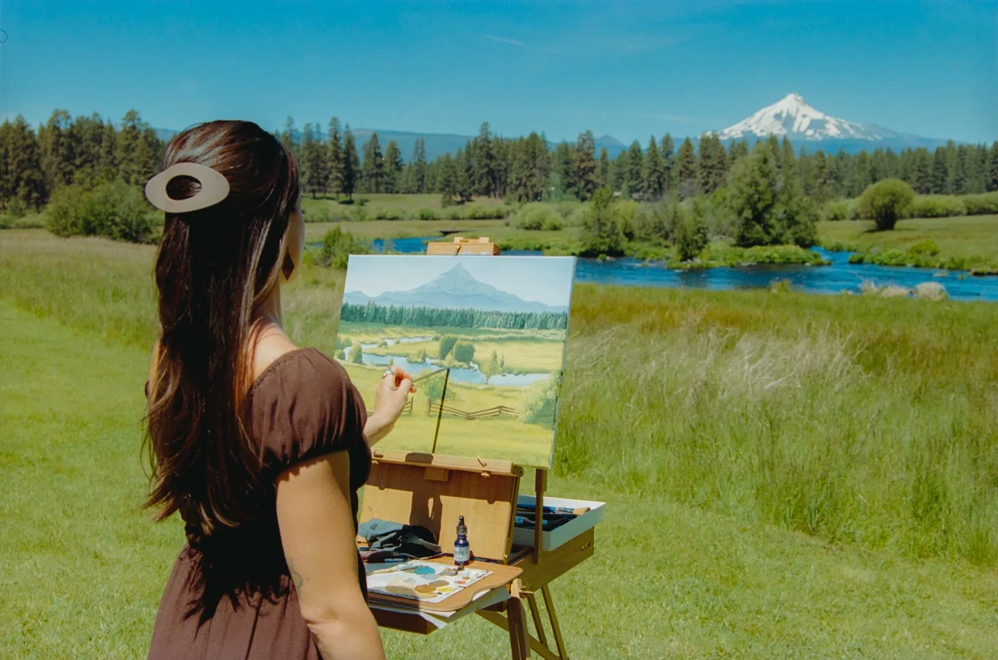 Holly Di Cecco painting live at an easel, mountain and river scene in the background